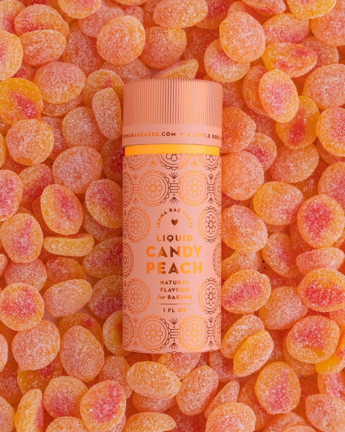 A cylindrical Liquid Candy Peach container from Jenna Rae Cakes rests on orange and yellow peach-shaped gummy candies, featuring a peach-colored design with floral patterns.