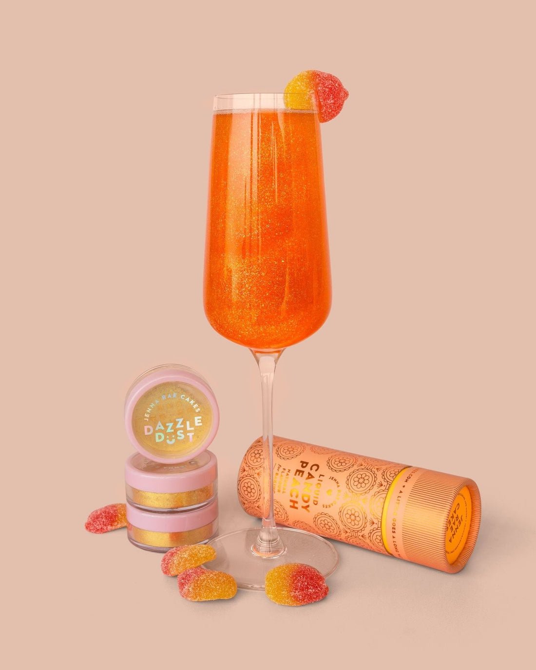 A Jenna Rae Cakes Liquid Candy Peach sparkling orange drink is served in a champagne flute with gummy candies inside and on the rim, next to “Dazzle Dust” containers and a cylindrical package, all set on a peach-colored background.
