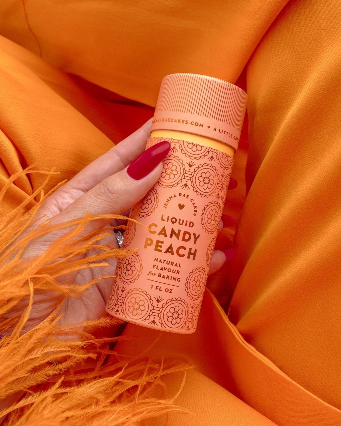 A hand with red nail polish holds a peach-colored Jenna Rae Cakes Liquid Candy Peach bottle, featuring a floral design, against vibrant orange fabric. The product is specially made for baking.