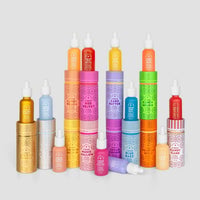 Bottles and cylindrical boxes of Jenna Rae Cakes’ Liquid Golden Bubbly skincare serum, each with vibrant colors and playful patterns indicating different scents, are arranged in rows on a white background.