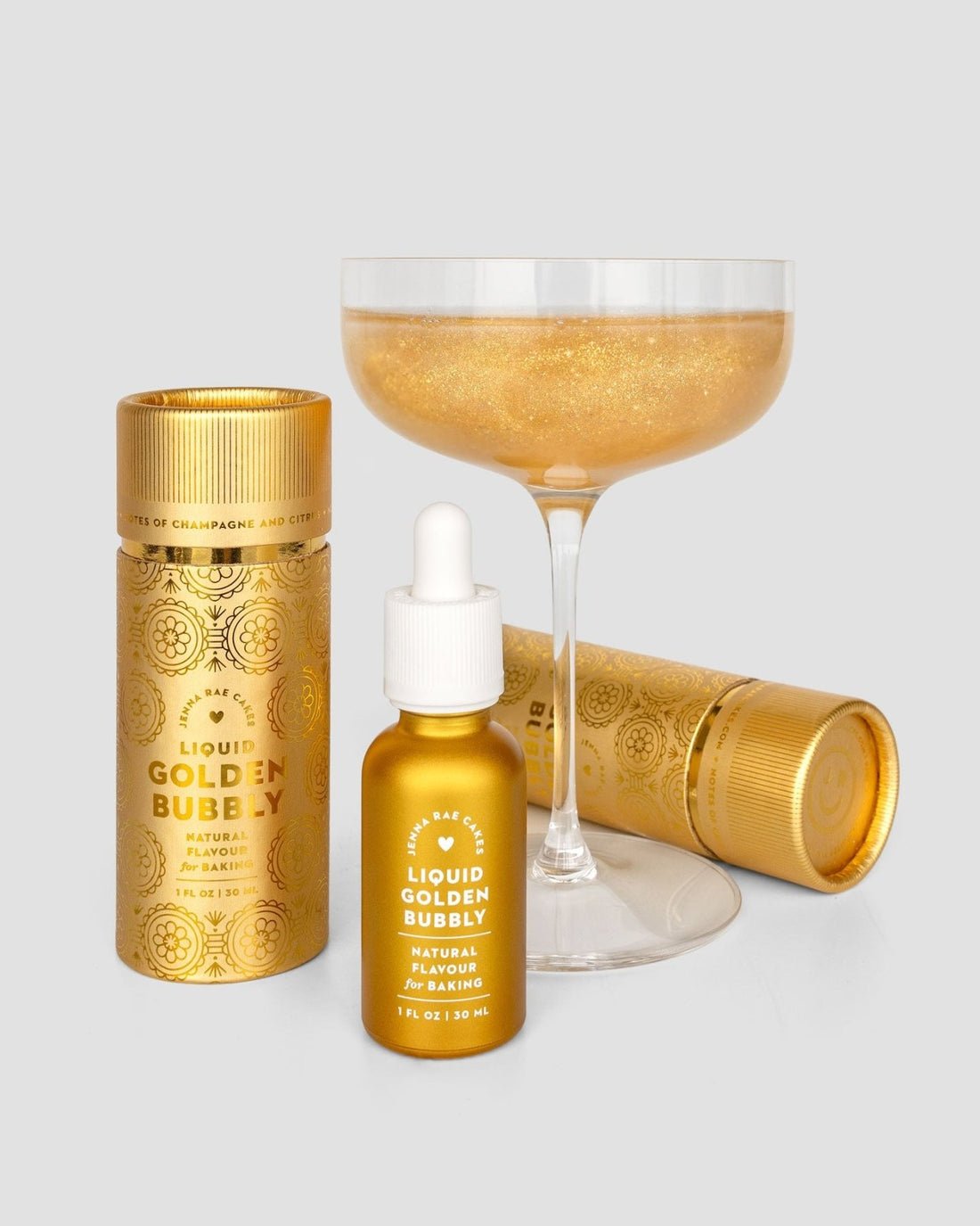 A coupe glass of sparkling golden liquid sits behind a dropper bottle and two cylindrical containers of Jenna Rae Cakes’ Liquid Golden Bubbly Natural Flavor for Baking in shiny gold packaging.