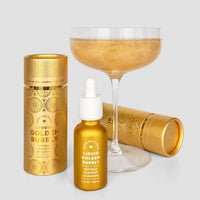 A champagne coupe of sparkling gold sits behind a gold dropper bottle and two gold cylinders labeled Liquid Golden Bubbly - Package of 6 by Jenna Rae Cakes, a natural flavour perfect for baking.