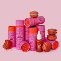 Jenna Rae Cakes’ Liquid Red Velvet - Package of 6 is displayed in pink and red cylindrical containers alongside stacked and broken red macarons and a small dropper bottle, all set against a pink background.