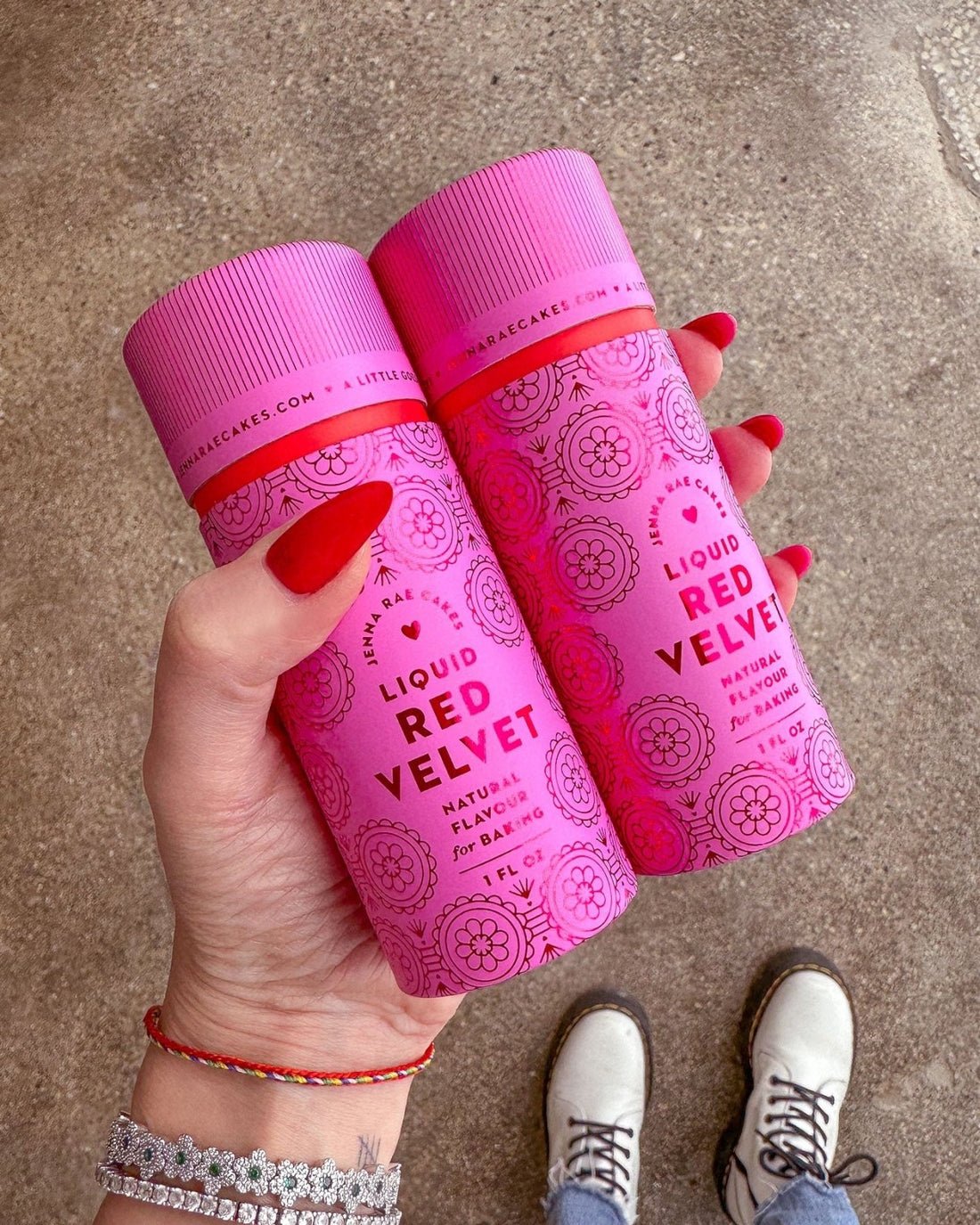A hand with red-painted nails holds two cans of Jenna Rae Cakes Liquid Red Velvet (Package of 6) against a concrete background. The person wears white shoes and beaded bracelets.
