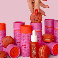 A hand with red nails holds a red macaron above a set of pink and red Jenna Rae Cakes Liquid Red Velvet - Package of 6 bottles and matching macarons, all arranged on a pink background.