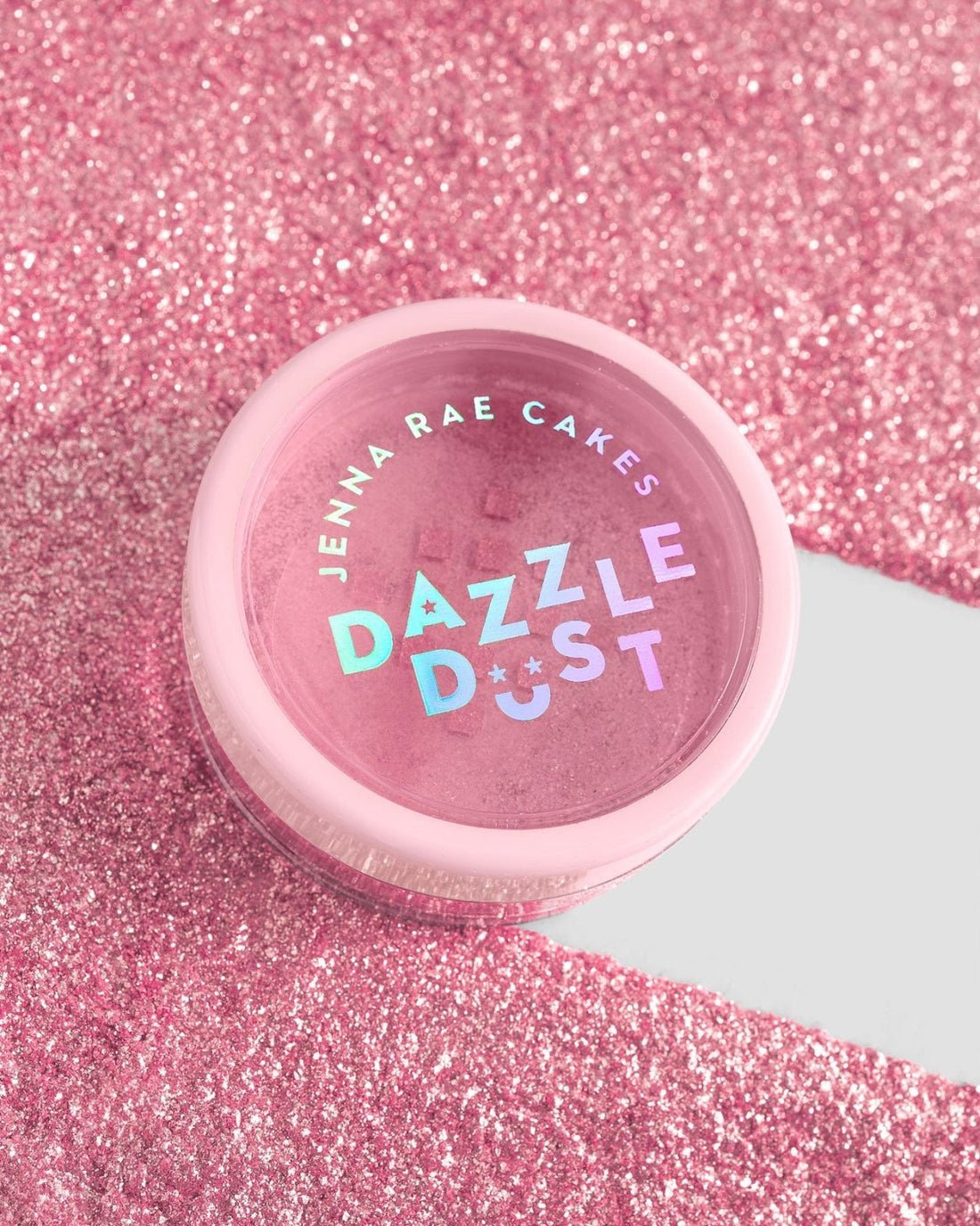 A small pink container labeled Jenna Rae Cakes Lollipop Edible Glitter - 5g Shaker (Package of 6) sits on a sparkling pink glitter surface, with white and iridescent text standing out on the lid.