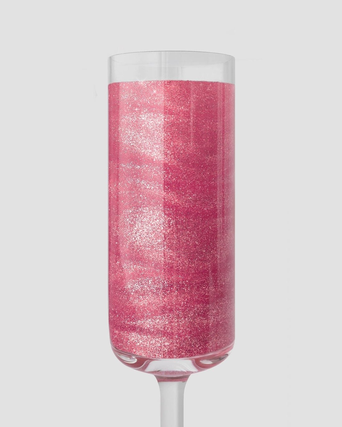 A clear champagne flute holds a vibrant pink drink sparkling with Jenna Rae Cakes Lollipop Edible Glitter, packaged in 5g shakers (set of 6), set against a plain light gray background.