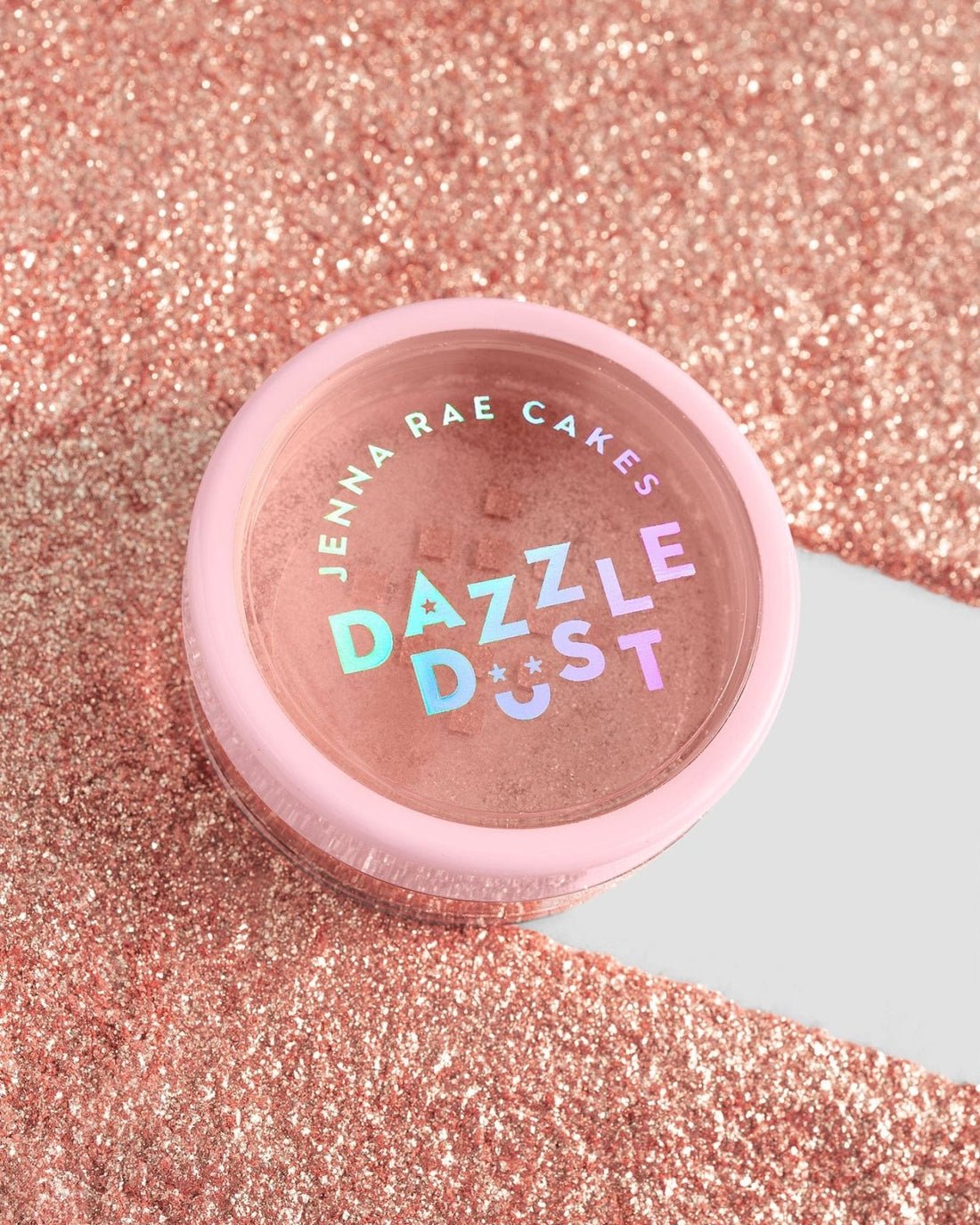 A package of six Jenna Rae Cakes Lucky Penny Edible Glitter 5g shakers, with pink lids and holographic labels, sits on a sparkling pink glitter surface reflecting various colors.