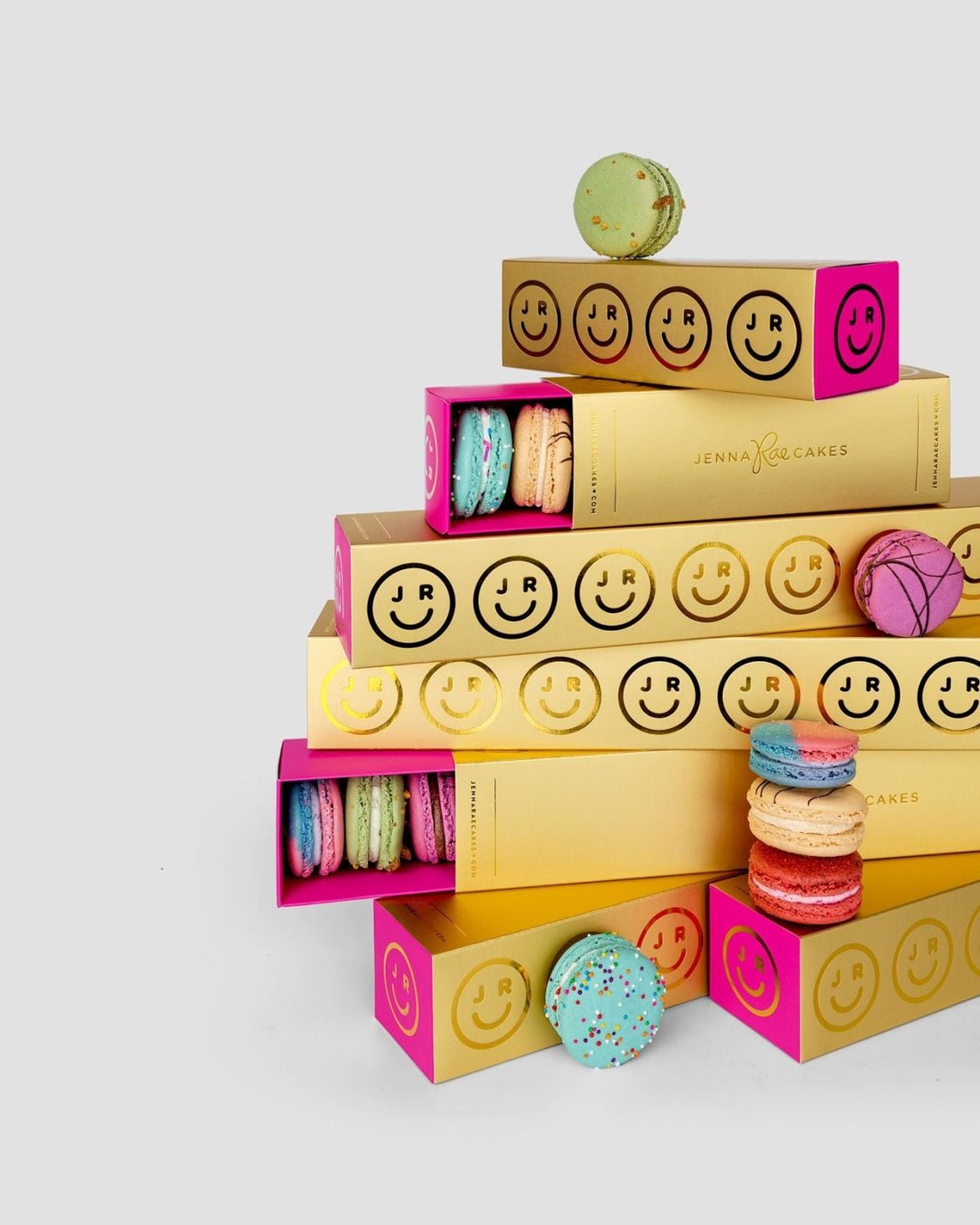 A stack of Jenna Rae Cakes Macaron Gift Boxes, adorned with smiley faces, is surrounded by colorful macarons against a plain light gray background.