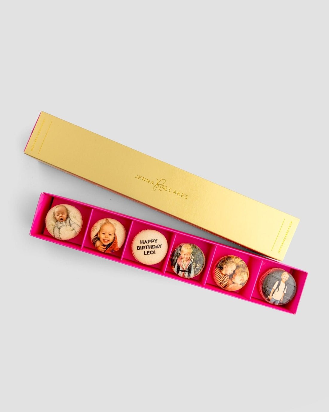The Macaron Message Gift Box by Jenna Rae Cakes is a rectangular gold box with six round macarons, each topped with a custom photo or message like Happy Birthday Leon! The bright pink interior adds a festive touch.