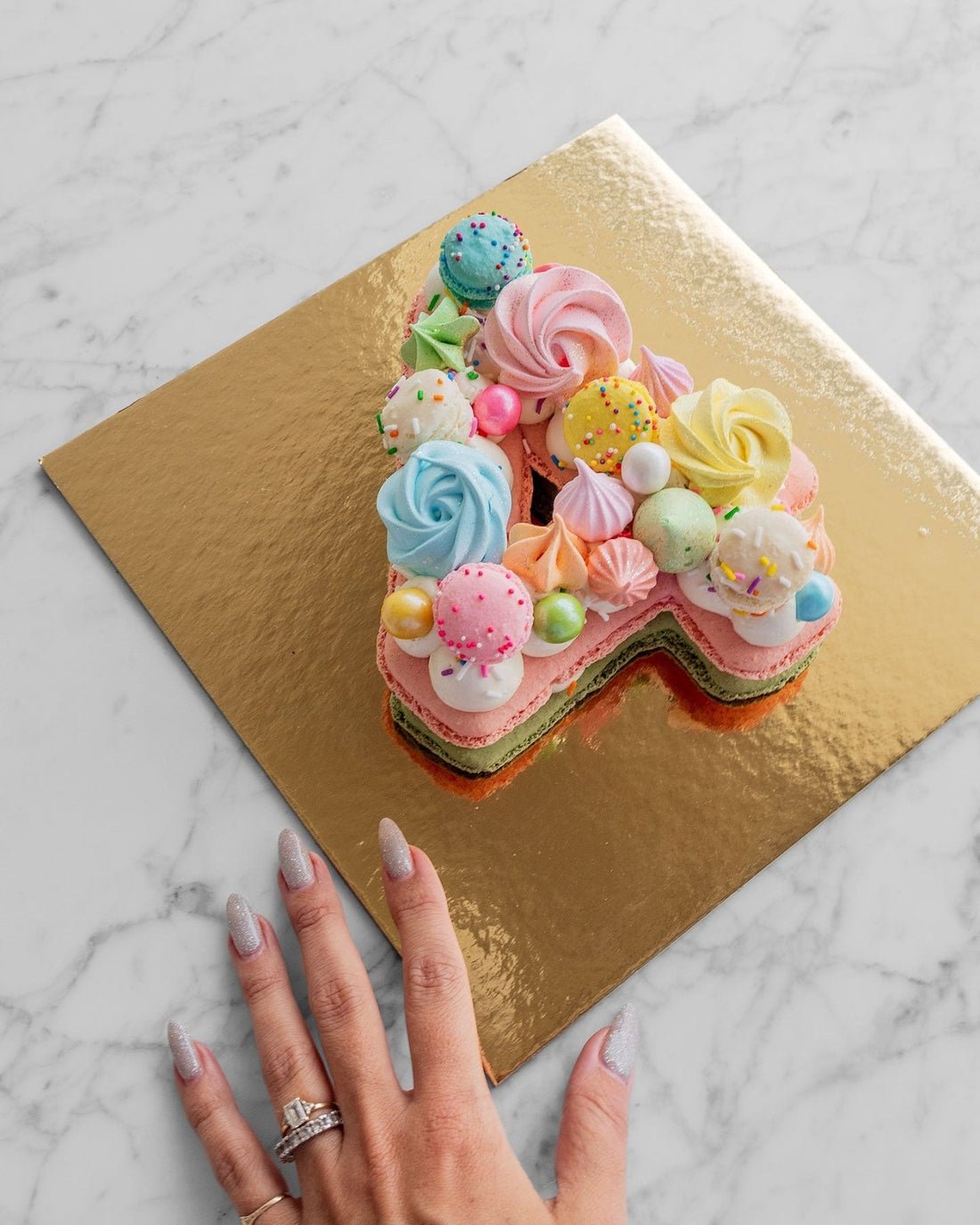A manicured hand reaches for a Macaron Number Cake by Jenna Rae Cakes, shaped like a colorful letter A and decorated with pastel meringues and sprinkles on a gold board atop marble.