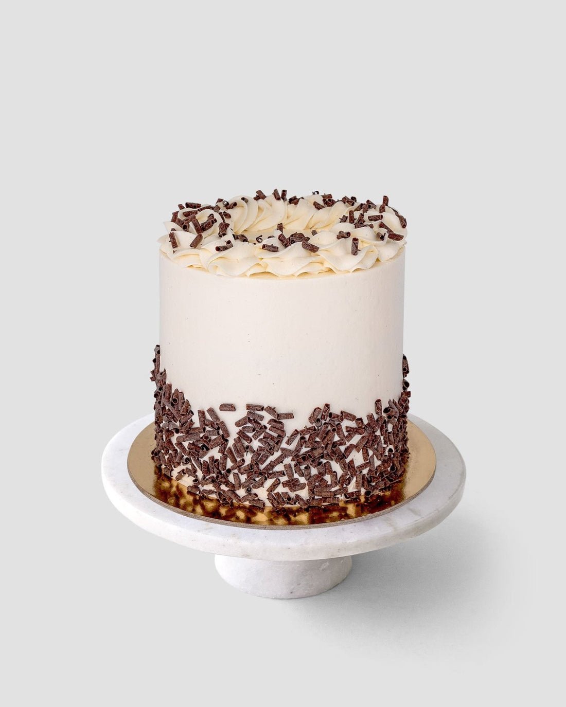 The Marble Cake by Jenna Rae Cakes is a tall, round cake with smooth white frosting, topped with icing swirls and chocolate shavings around the base, displayed on a white marble cake stand against a plain background.