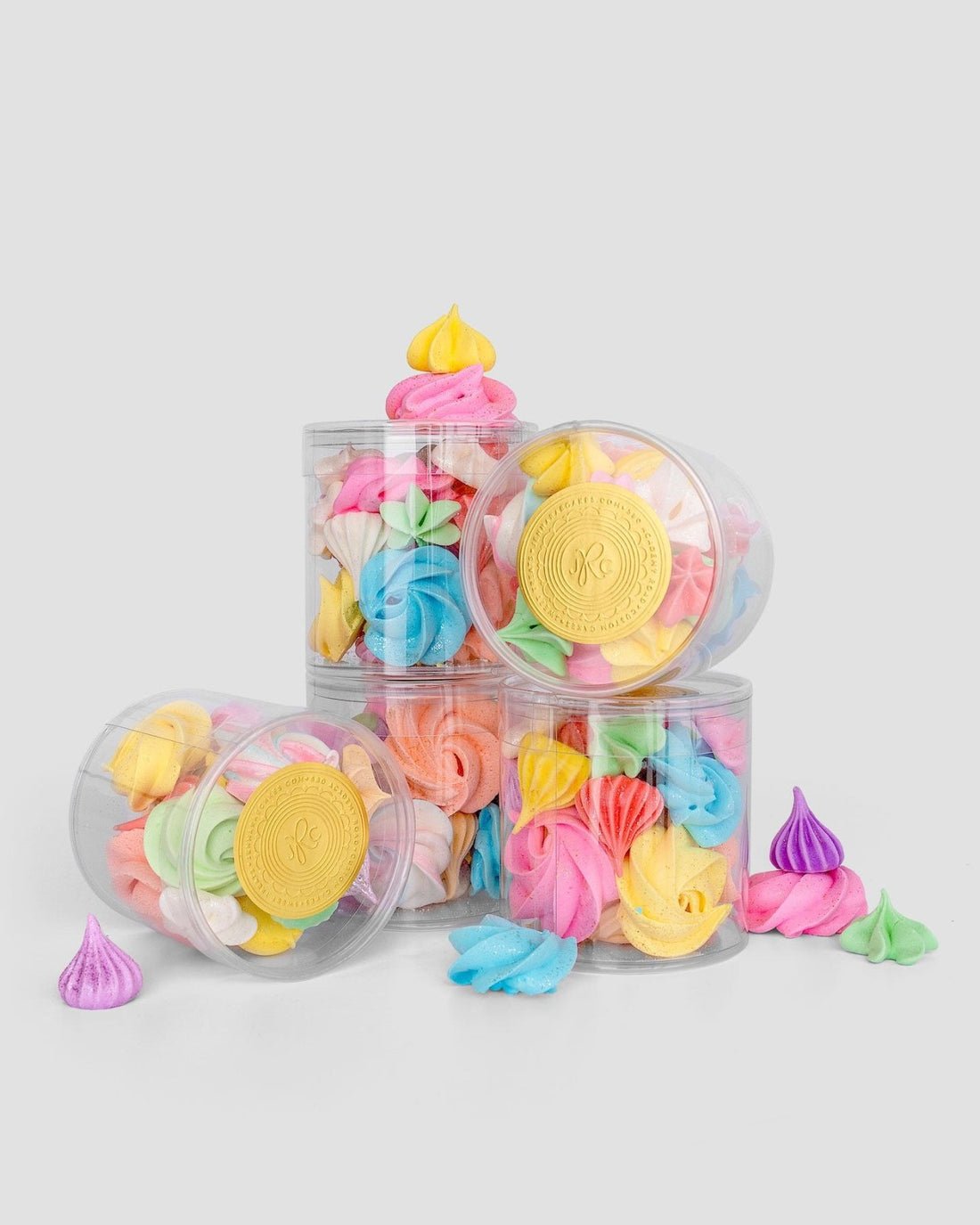 Jenna Rae Cakes Meringues in clear plastic jars feature colorful cookies in pastel pink, blue, yellow, green, and purple, stacked on a gray background with a few meringues scattered around.