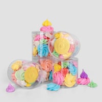 Jenna Rae Cakes Meringues in clear plastic jars feature colorful cookies in pastel pink, blue, yellow, green, and purple, stacked on a gray background with a few meringues scattered around.