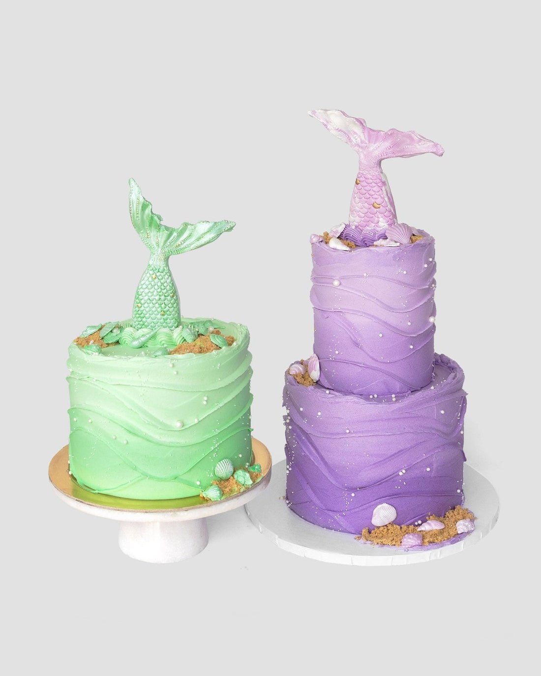 Mermaid Cakes by Jenna Rae Cakes: one green, single-tiered and one tall, purple, two-tiered—both topped with mermaid tails and decorated with wave-like frosting, seashells, and edible pearls.