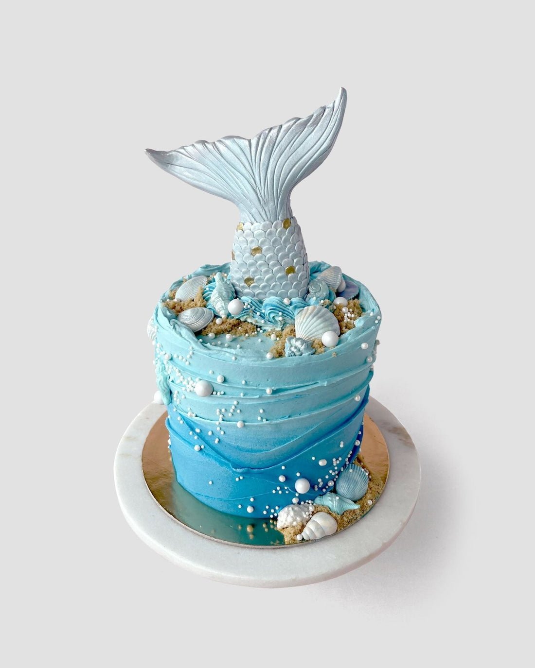 The Mermaid Cake by Jenna Rae Cakes is a round blue cake adorned with edible seashells, pearls, sand-like crumbs, and topped with a large silver mermaid tail. Presented on a white and gold cake board.