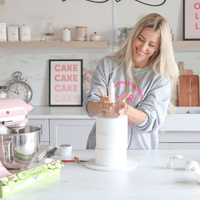 A woman smiles while decorating a tall white cake in a modern kitchen. Baking tools, a pink mixer, and cake supplies are on the counter, with Jenna Rae Cakes Online Classes visible on the wall behind her.