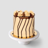 The Peanut Butter Cup Cake by Jenna Rae Cakes is a tall, round cake with light brown frosting, chocolate drip, and swirls of frosting and chocolate pieces on top, displayed on a white marble cake stand against a gray background.