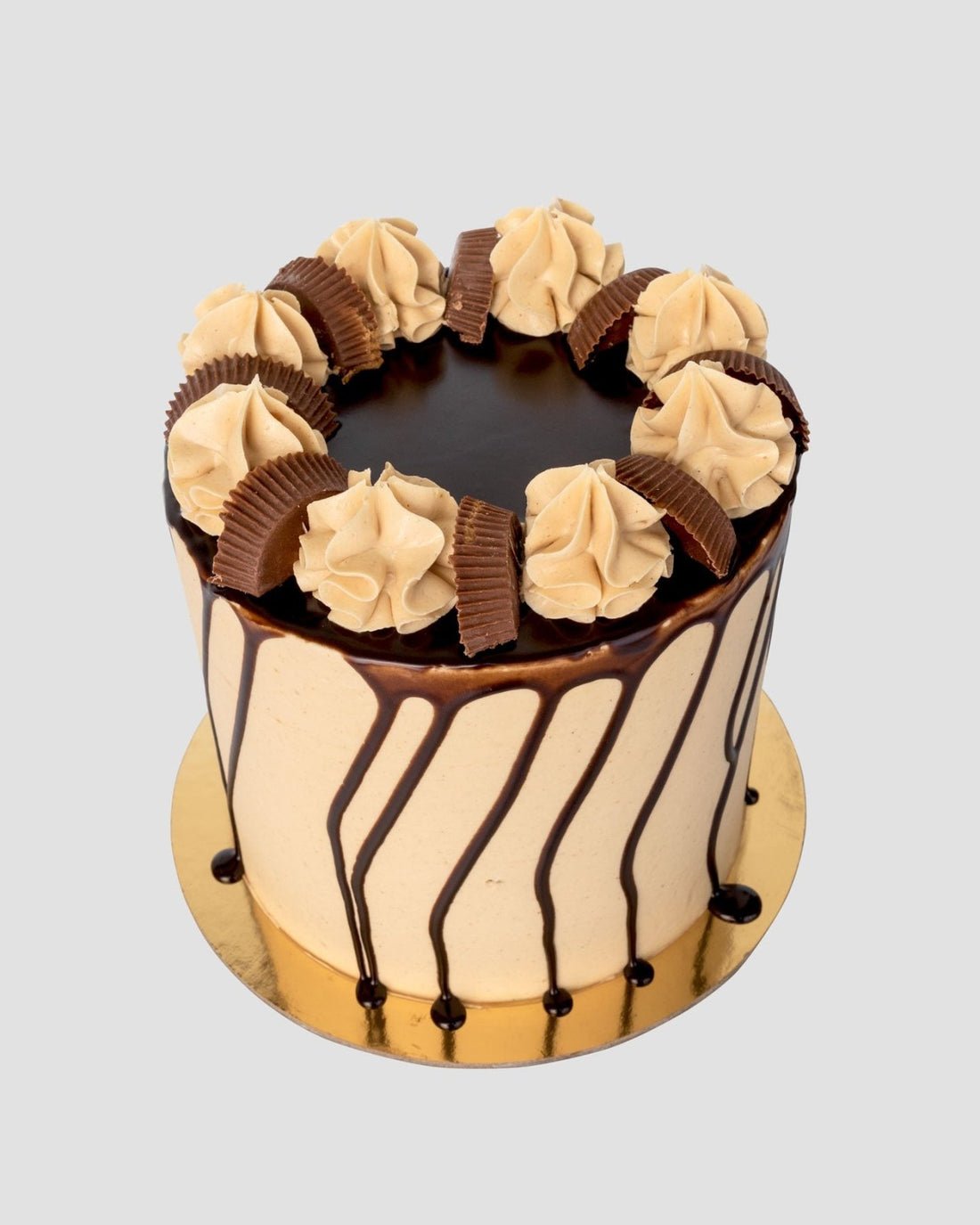 The Peanut Butter Cup Cake by Jenna Rae Cakes is a tall cake with light brown frosting, chocolate drip, swirls of frosting, and halved chocolate peanut butter cups on a gold cake board.