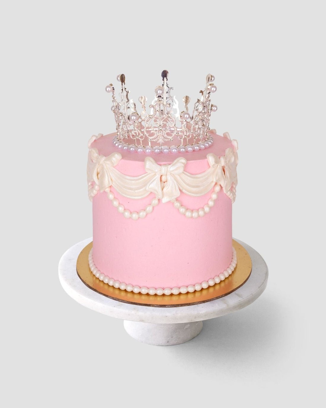 The Princess Crown Cake by Jenna Rae Cakes features pink frosting, white icing bows, pearly beads, and a decorative edible crown, presented on a round marble stand against a simple light background.