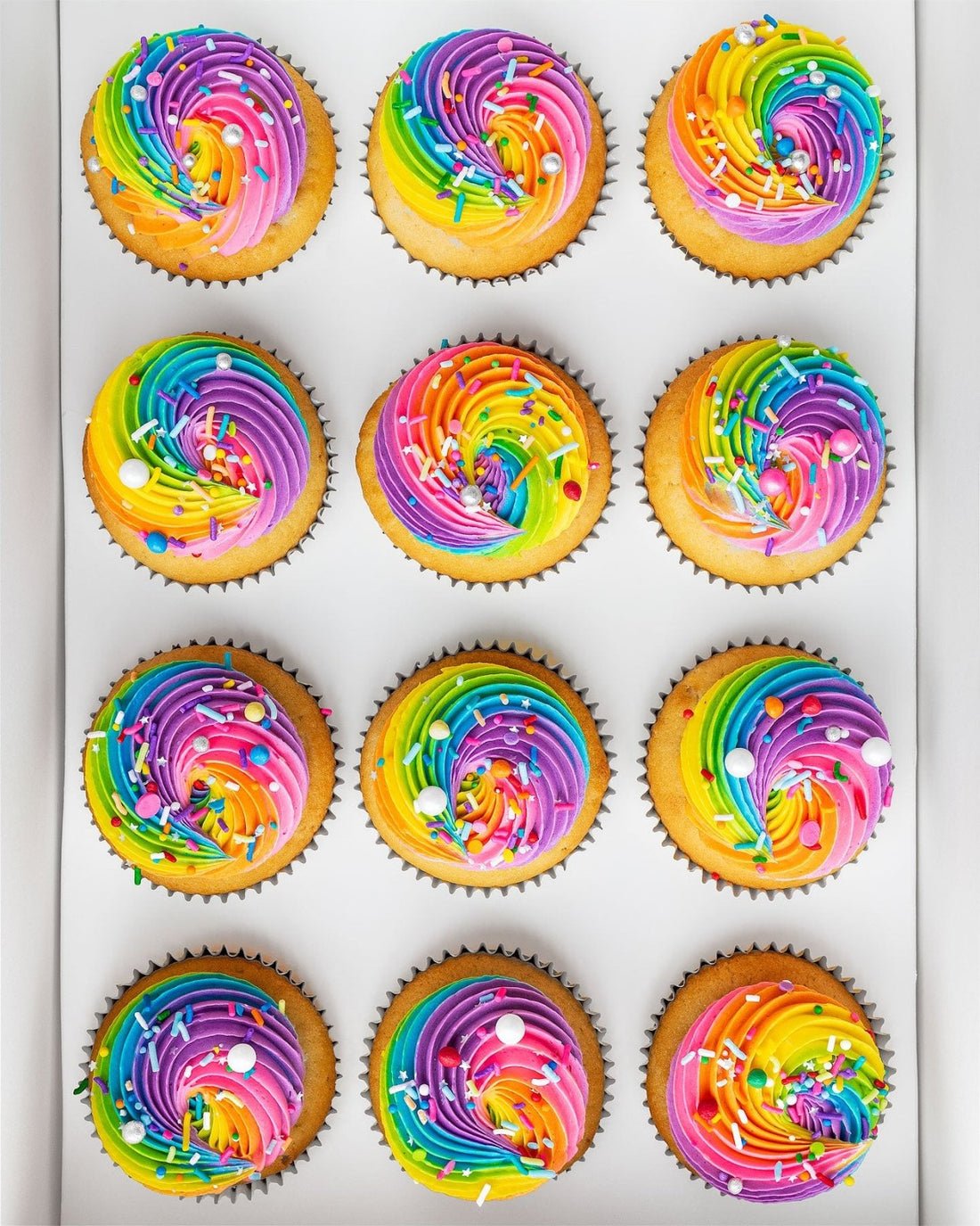 Rainbow Cupcakes by Jenna Rae Cakes: twelve cupcakes with vibrant rainbow swirled frosting and colorful sprinkles, arranged in three rows on a white tray.