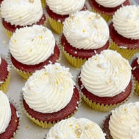 Rows of red velvet cupcakes with swirls of white frosting, topped with white and gold sprinkles, edible pearls, and glitter, arranged on a tray.