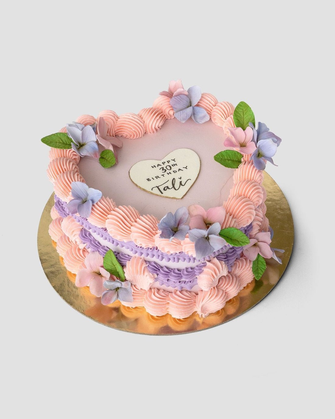 The Sugar Flower Heart Cake by Jenna Rae Cakes features pink and purple frosting, pastel flowers, green leaves, and a heart-shaped cookie with Happy 30th Birthday Tali, all presented on a gold board.
