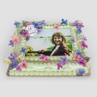 The Sugar Flower Sheet Cake by Jenna Rae Cakes is a rectangular cake adorned with pink, purple, and blue sugar flowers, piped cream borders, and an edible outdoor photo of a smiling woman with a 60 & proud topper.