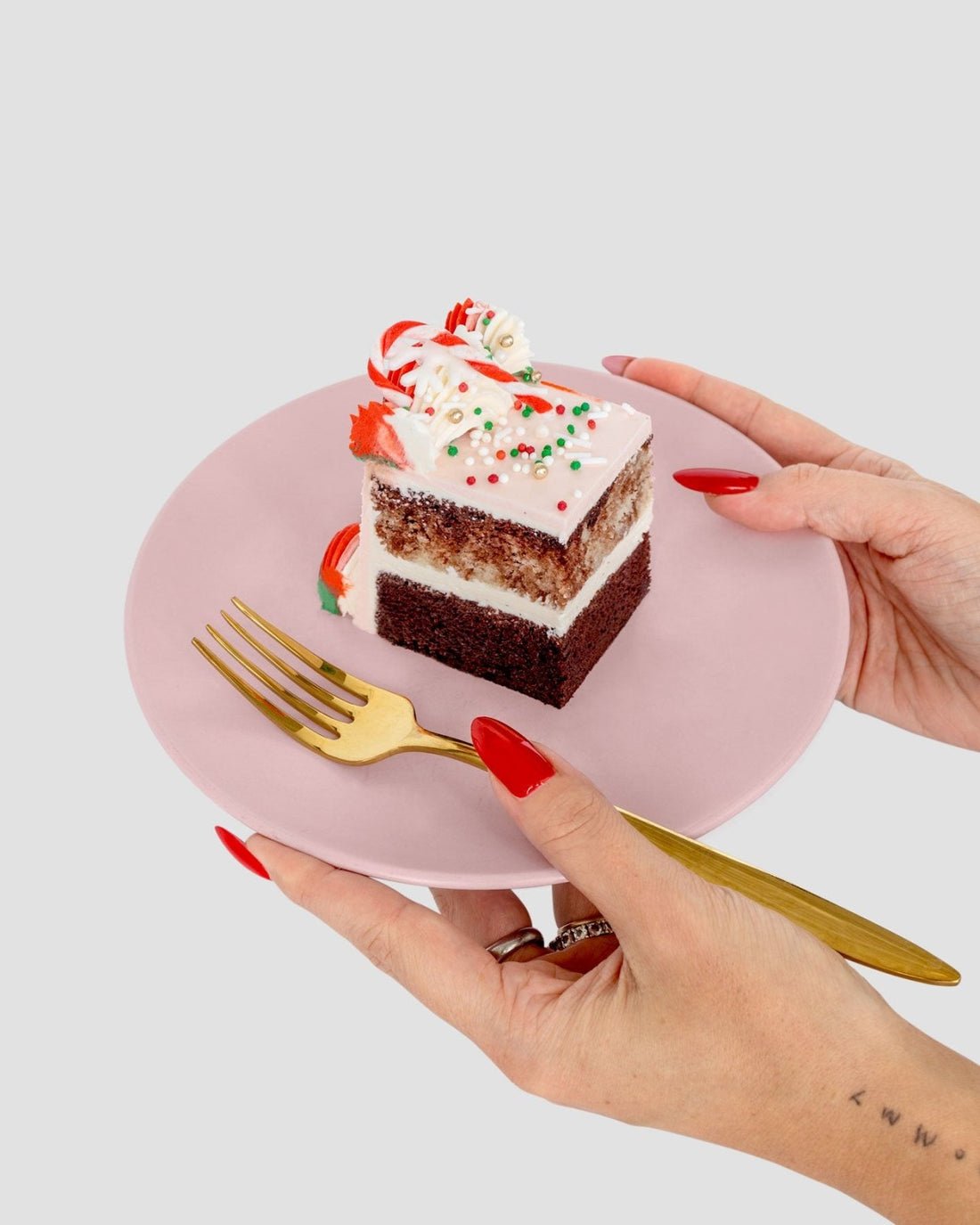 A person with red nails holds a pink plate with a slice of Jenna Rae Cakes’ Sugar Flower Sheet Cake, topped with white frosting, sprinkles, and candy, while holding a gold fork in the other hand.