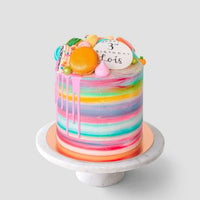 The Half Works Cake by Jenna Rae Cakes features a tall, colorful watercolor design with pink drip icing, topped with assorted macarons, meringues, and a white disc reading 3rd birthday Boris, displayed on a marble cake stand.
