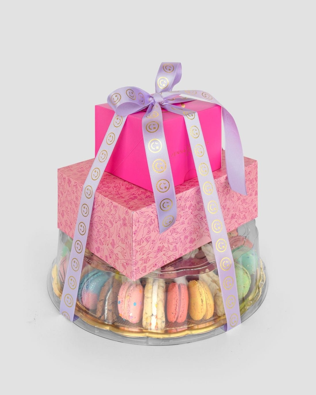 The Jenna Rae Cakes Treat Tower features two pink gift boxes tied with a lavender ribbon, placed on a clear tray filled with assorted colorful macarons, set against a plain light gray background.