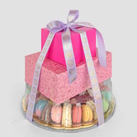 The Jenna Rae Cakes Treat Tower features two pink gift boxes tied with a lavender ribbon, placed on a clear tray filled with assorted colorful macarons, set against a plain light gray background.