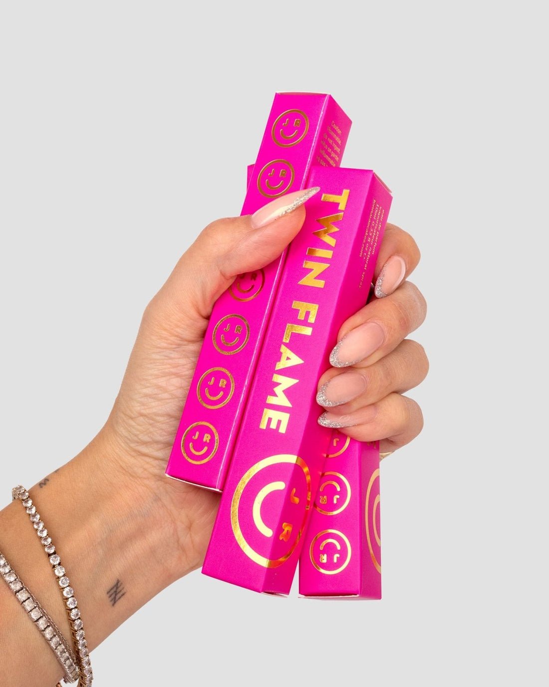 A hand with manicured nails holds two bright pink Twin Flame Perfume boxes from Jenna Rae Cakes, featuring gold lettering and smiley face designs; the wearer has a beaded bracelet and a small wrist tattoo.