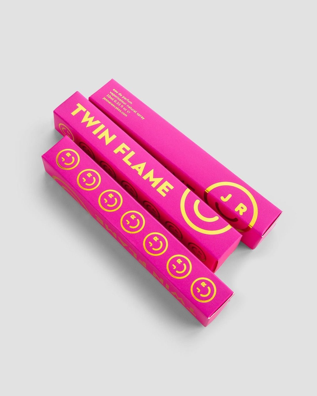 Two bright pink rectangular boxes labeled Twin Flame Perfume in bold yellow text, featuring yellow smiley faces and J R in a circle, from Jenna Rae Cakes, displayed on a light gray background.