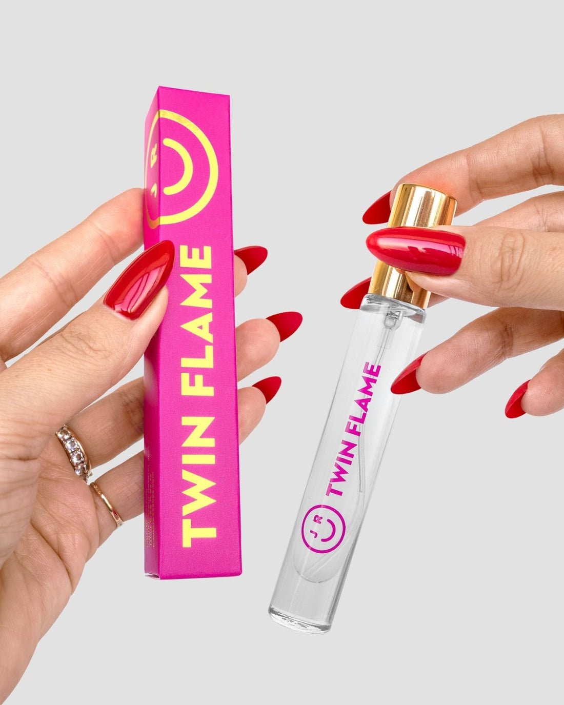 A person with red nails holds a pink Twin Flame Perfume box and a clear Jenna Rae Cakes Twin Flame Perfume bottle with a gold cap, against a light gray background.