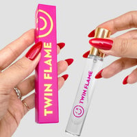 A person with red nails holds a pink Twin Flame Perfume box and a clear Jenna Rae Cakes Twin Flame Perfume bottle with a gold cap, against a light gray background.