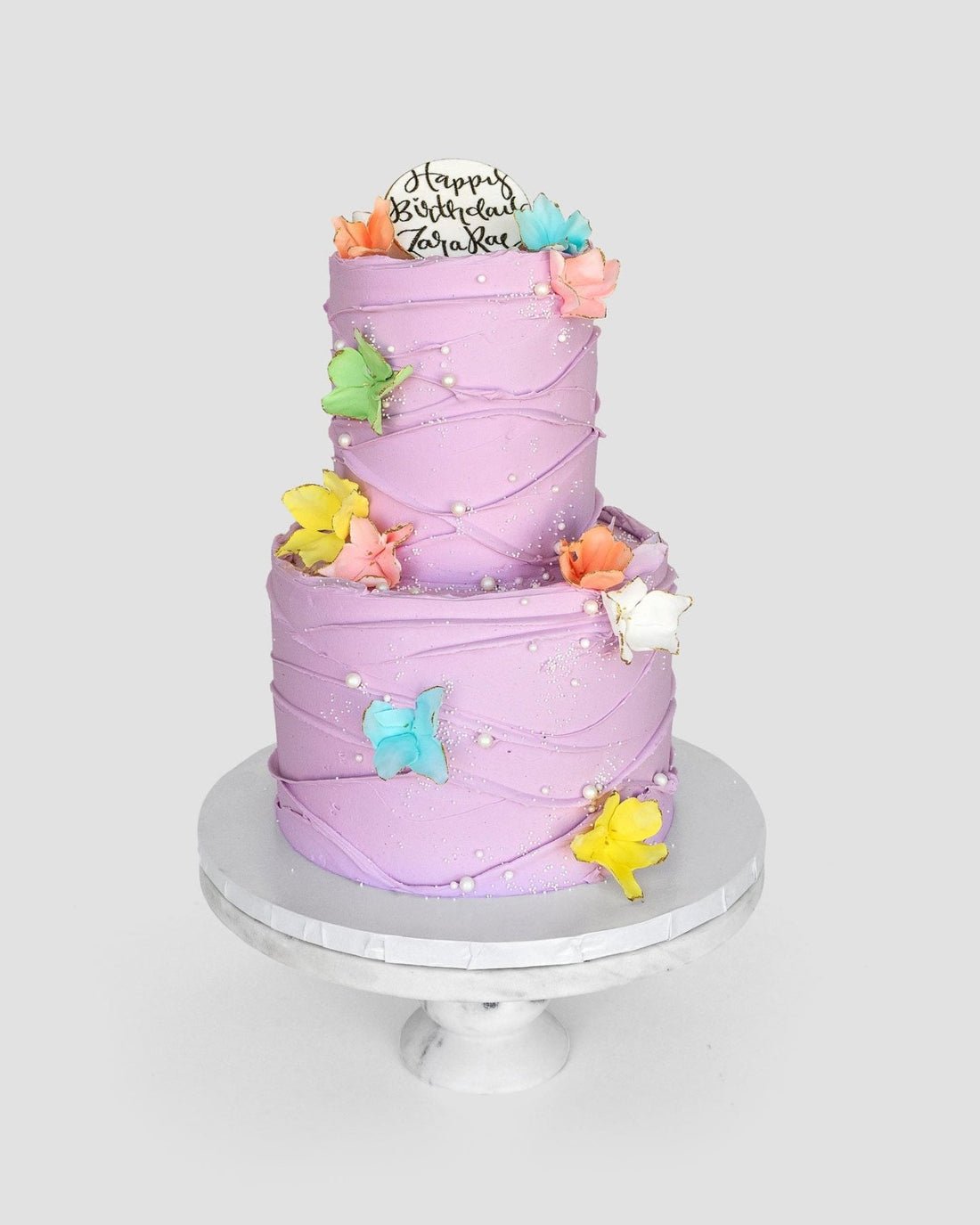 The Waves and Pearls cake by Jenna Rae Cakes is a two-tiered lavender creation adorned with colorful edible butterflies, white pearls, and a “Happy Birthday Zariah” topper, presented on a white cake stand.