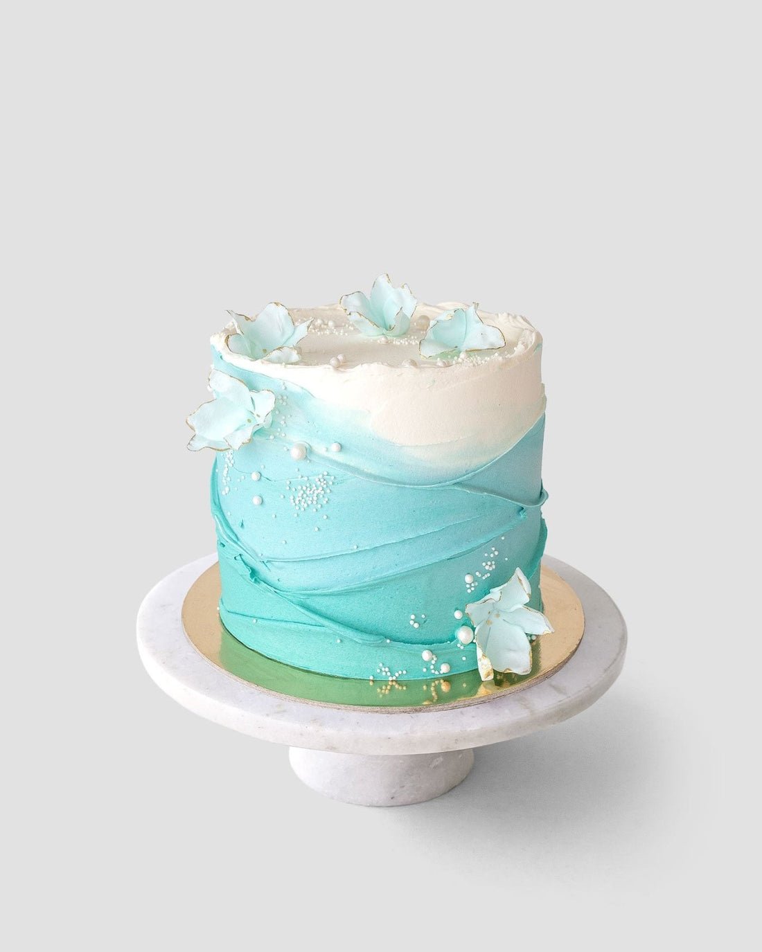 The Waves and Pearls cake by Jenna Rae Cakes features blue and white ombre textured icing, adorned with edible blue and white flowers and pearl-like sprinkles, displayed on a marble stand against a light gray background.