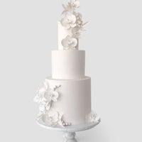 A three-tiered white cake from Jenna Rae Cakes Wedding Cakes Under $1,000 collection features elegant sugar flowers and leaves, displayed on a pedestal cake stand against a light gray background.