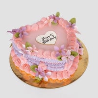 Jenna Rae Cakes’ Wedding Cakes Under $200 features a heart-shaped design with pink and purple frosting, purple flowers, green leaves, and a white “Happy Birthday” topper on a gold board.