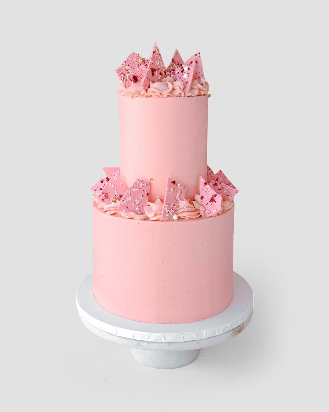 The Wedding Cakes Under $500 by Jenna Rae Cakes features a two-tiered pink cake with pink frosting swirls, pink chocolate shards, and sprinkles, shown on a white stand against a simple backdrop.