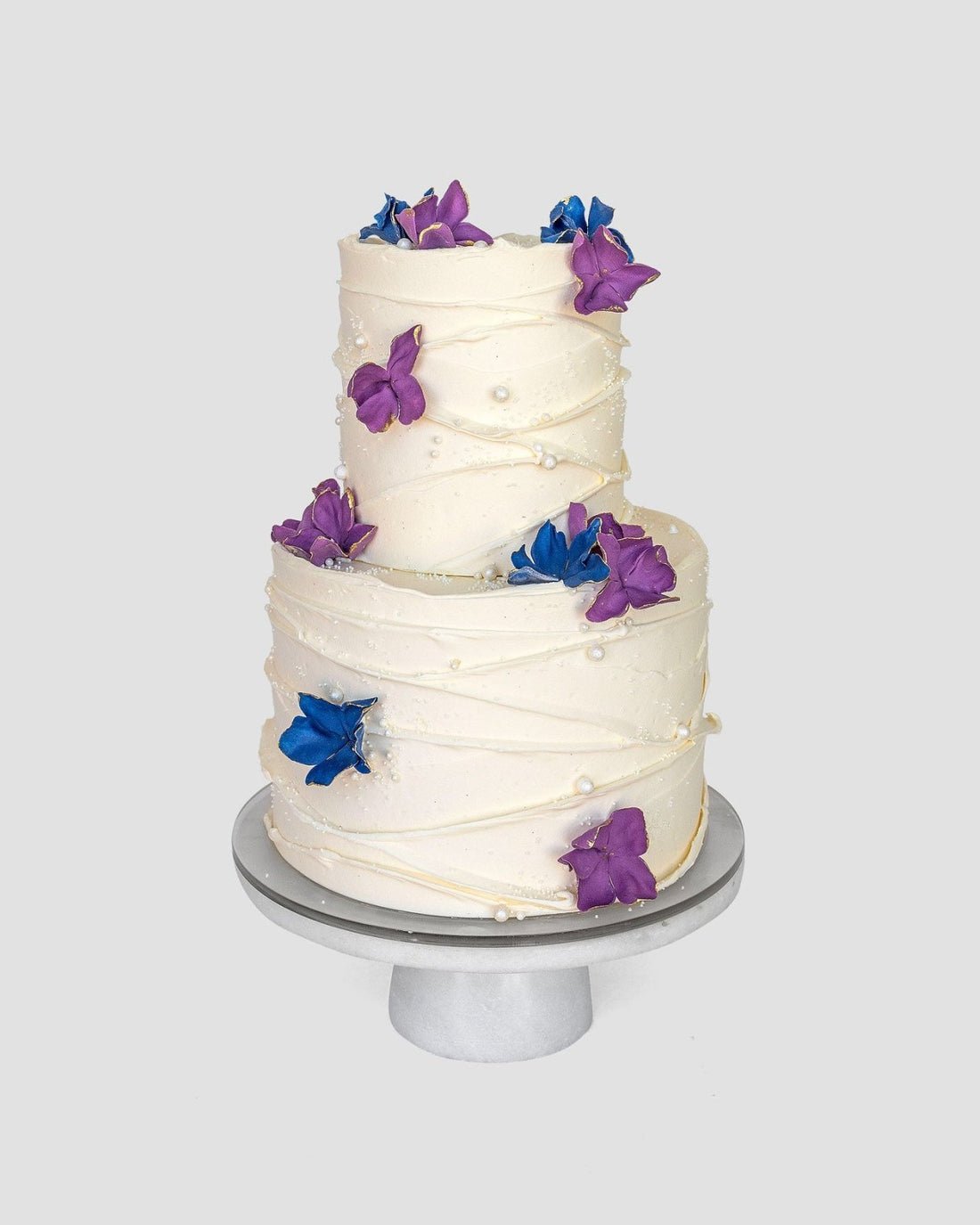 From Jenna Rae Cakes Wedding Cakes Under $500 collection: a two-tiered cake with white frosting, decorated with purple and blue flowers and pearl-like accents, displayed on a round white stand against a light gray background.