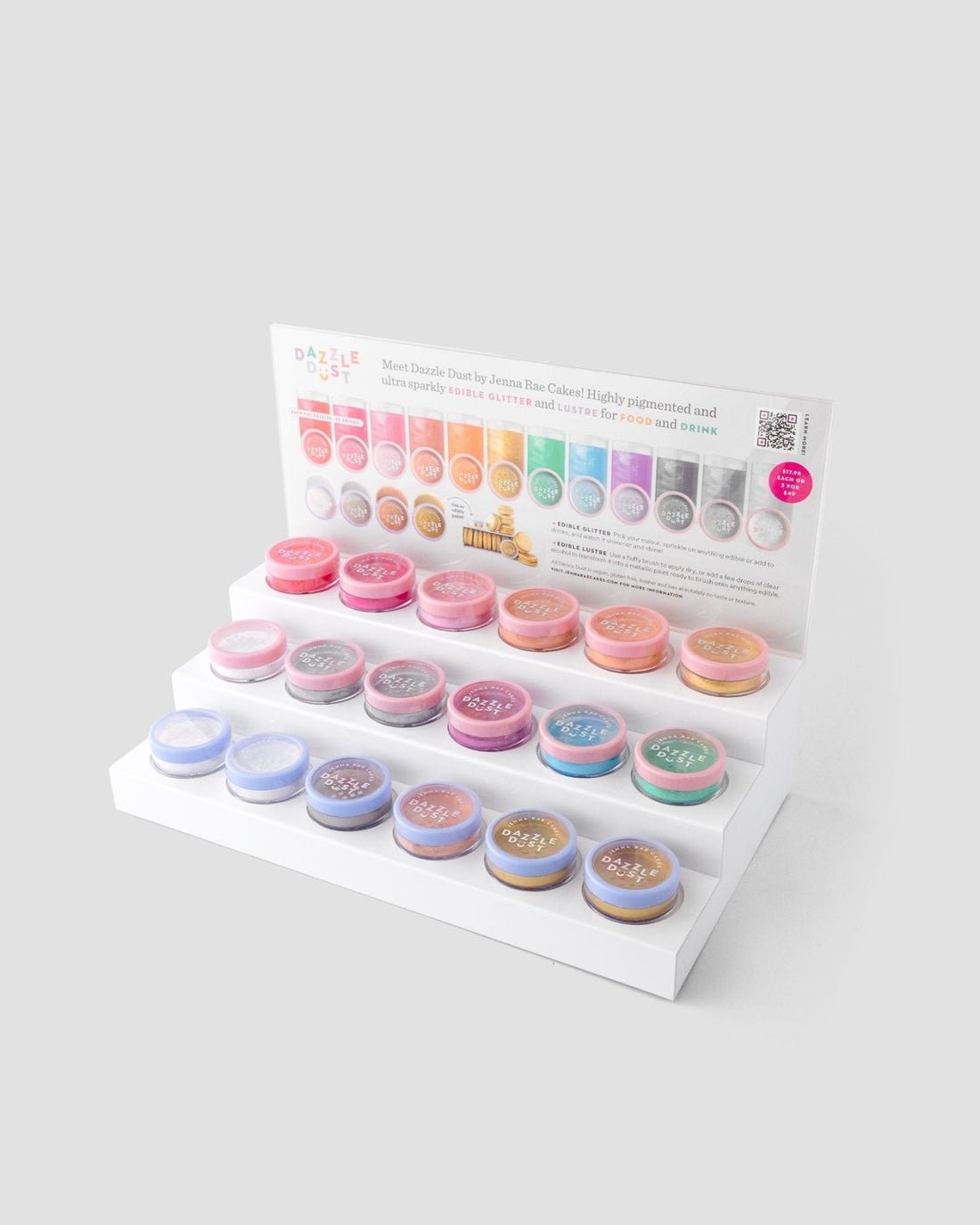 The White Acrylic Dazzle Dust Stand by Jenna Rae Cakes holds small round jars of shimmer powders, with three tiers and a sign at the back showing color options and product info. Package includes 1 stand.