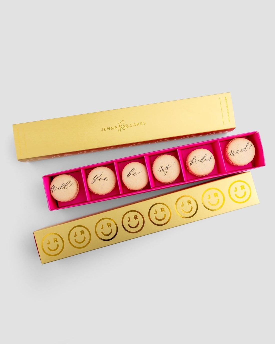The Jenna Rae Cakes Will You Be My... Gift Box features a gold box with six macarons spelling “Will you be my bridesmaid?” in cursive and a pink box of six macarons decorated with smiley faces and initials, set against a plain background.