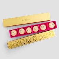 The Jenna Rae Cakes Will You Be My... Gift Box features a gold box with six macarons spelling “Will you be my bridesmaid?” in cursive and a pink box of six macarons decorated with smiley faces and initials, set against a plain background.