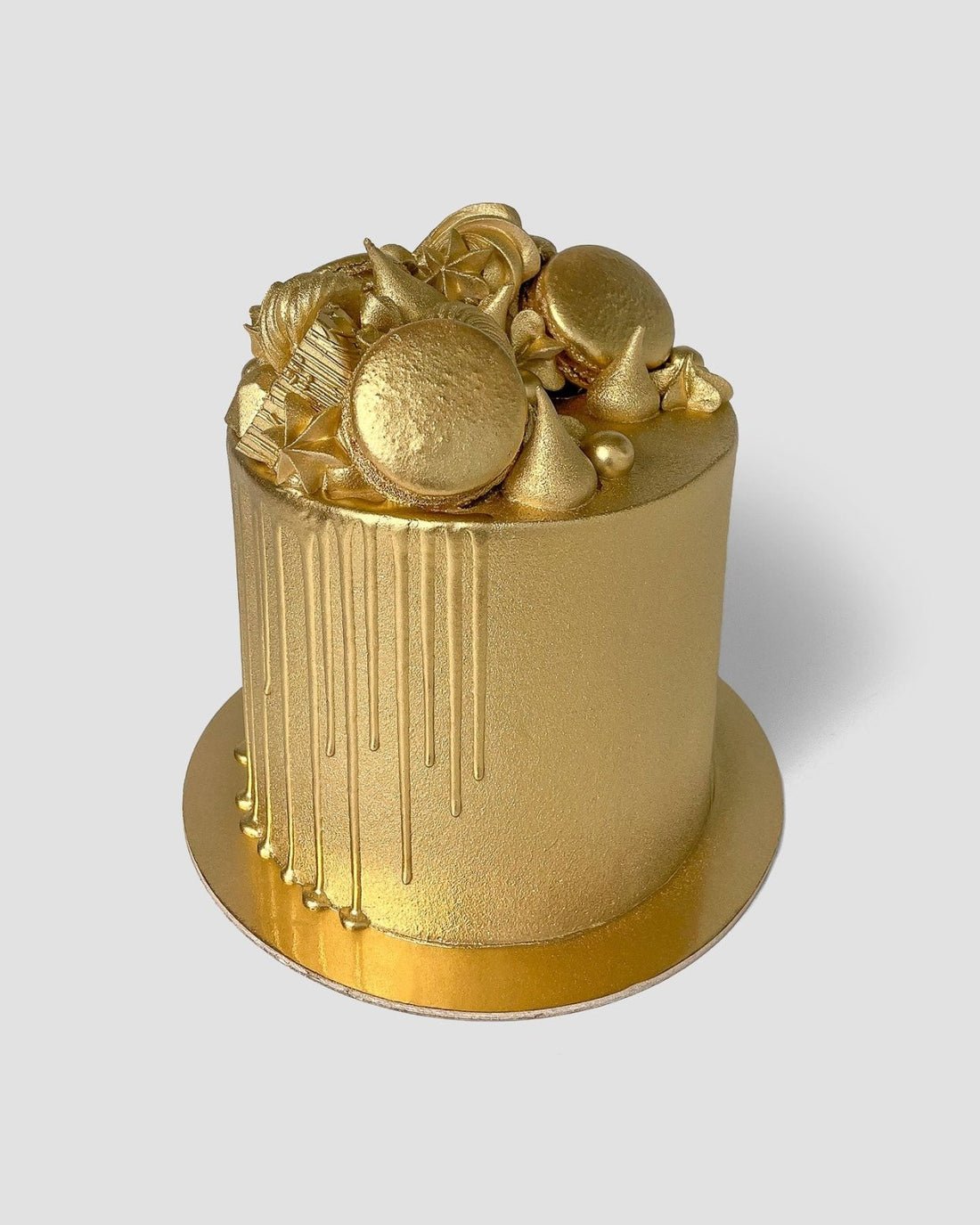 A round cake glistens with Jenna Rae Cakes’ Antique Gold Edible Lustre Dust, featuring metallic gold frosting, gold drips, macarons, candies, and decorations on a matching gold board set against a plain gray backdrop.