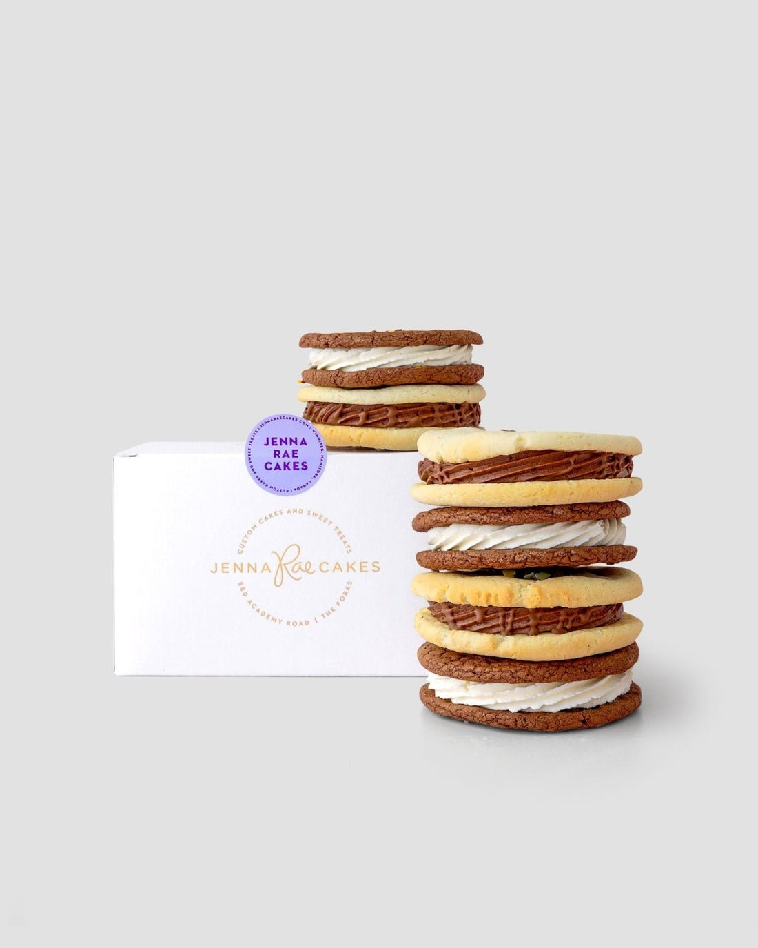A stack of cookie sandwiches from the Jenna Rae Cakes Cookie Sandwich Gift Box is displayed in front of a white box on a light grey background.