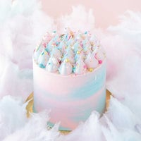 The Cotton Candy Cake by Jenna Rae Cakes features swirled pink and blue whipped frosting, colorful sprinkles, and fluffy cotton candy accents, set against a pale pink background.