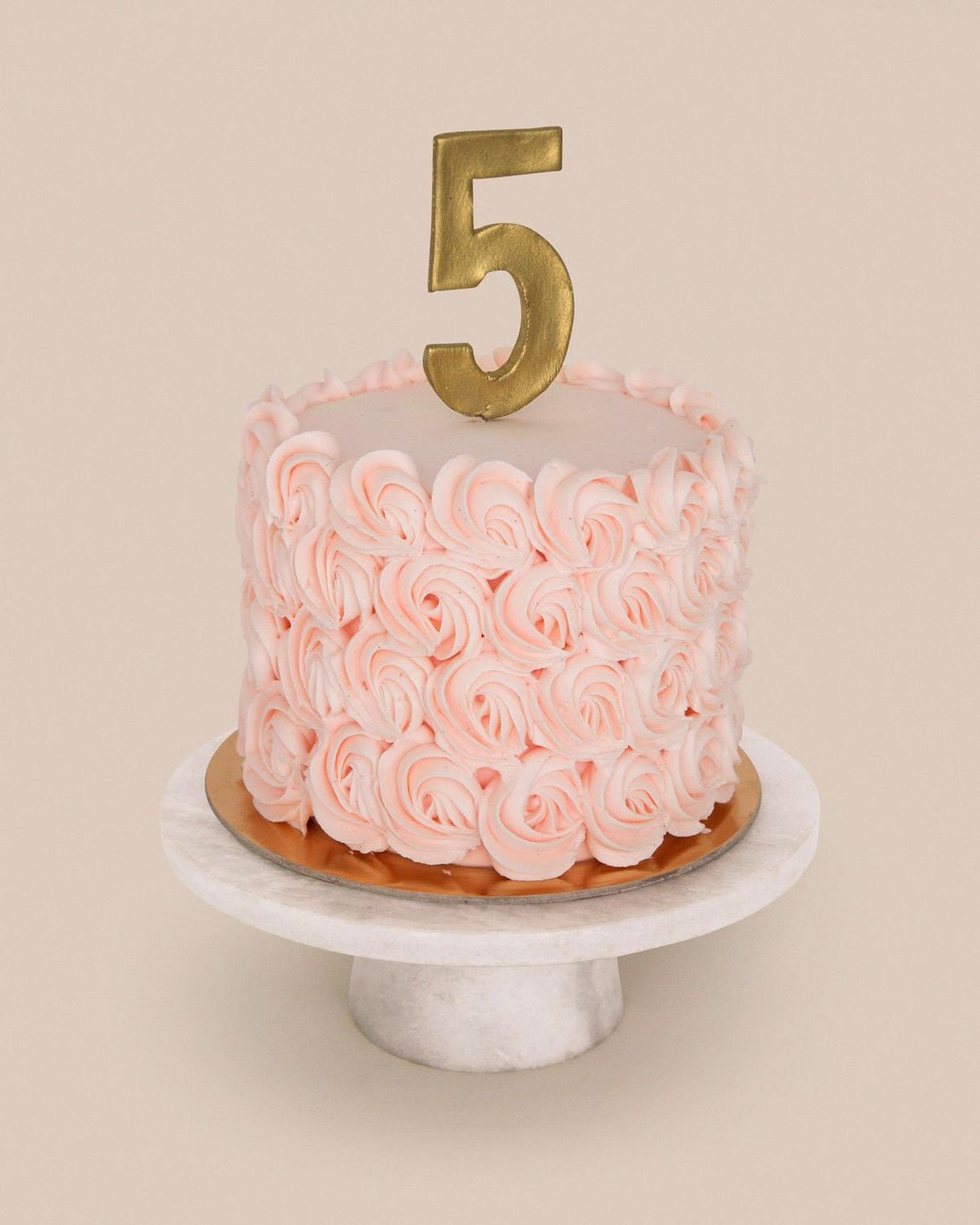 The Jenna Rae Cakes Design Your Own Cake is shown with pink rosettes, a gold number 5 candle, and displayed on a marble stand against a beige background.