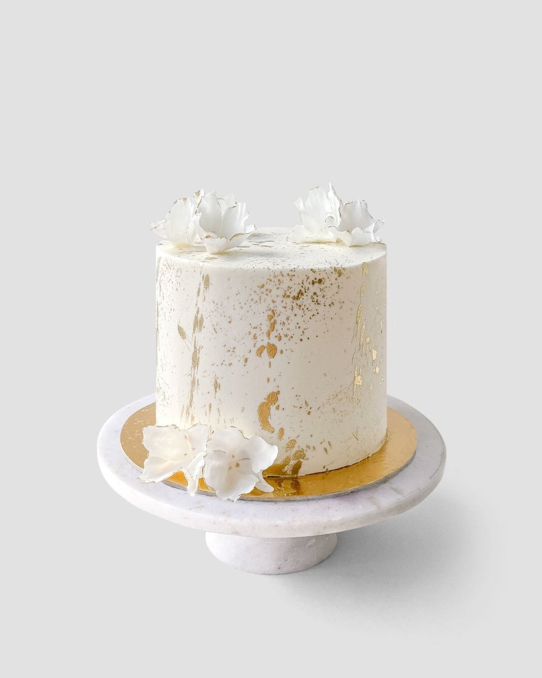 The Design Your Own Cake by Jenna Rae Cakes features white frosting with gold splatters and delicate white flowers, presented on a round marble stand against a light gray background.